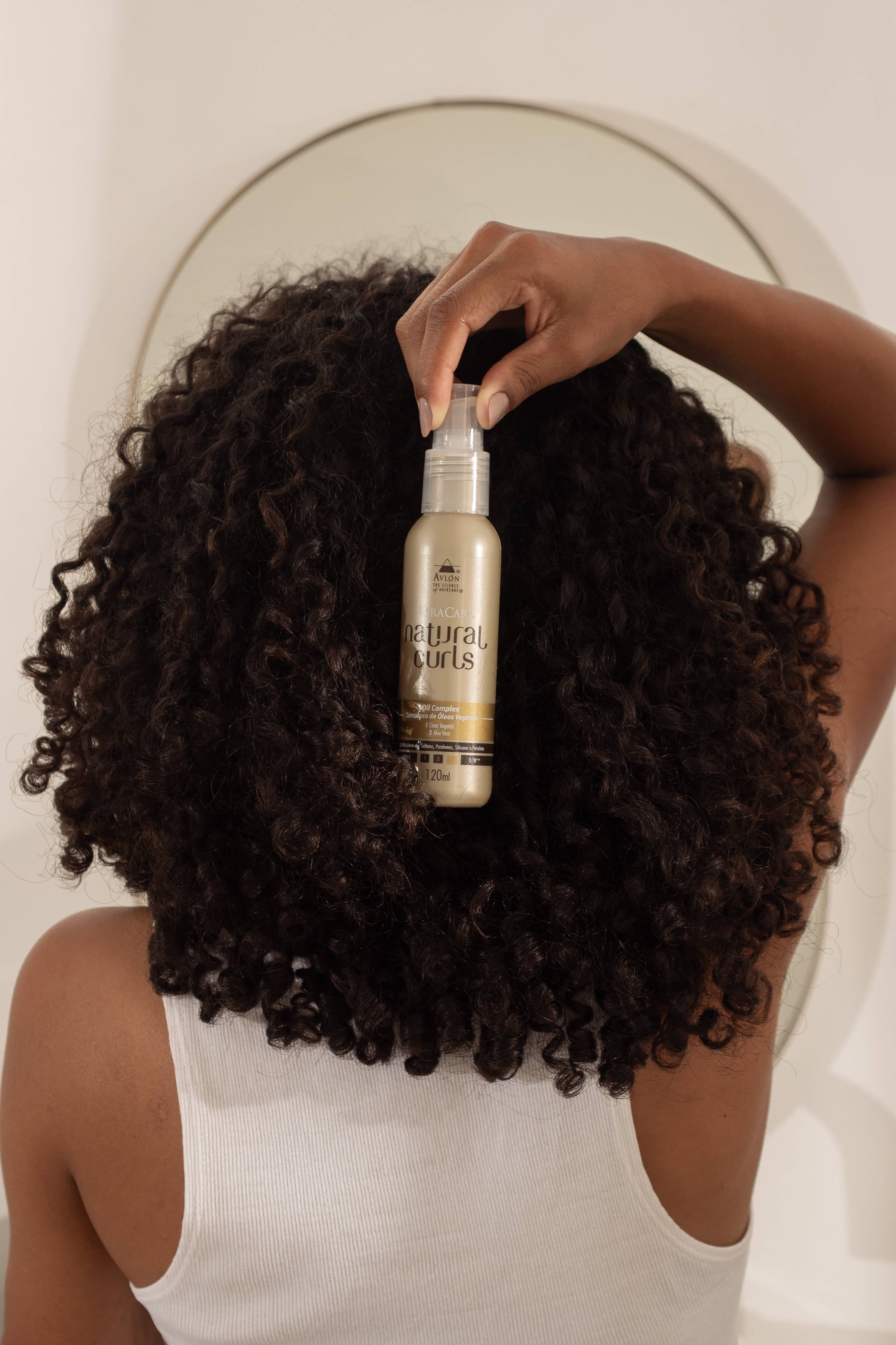 Mulher com cabelo cacheado segurando o Oil Complex Natural Curls em frente aos fios, mostrando óleo nutritivo para definição, brilho e umectação de cabelos ressecados.